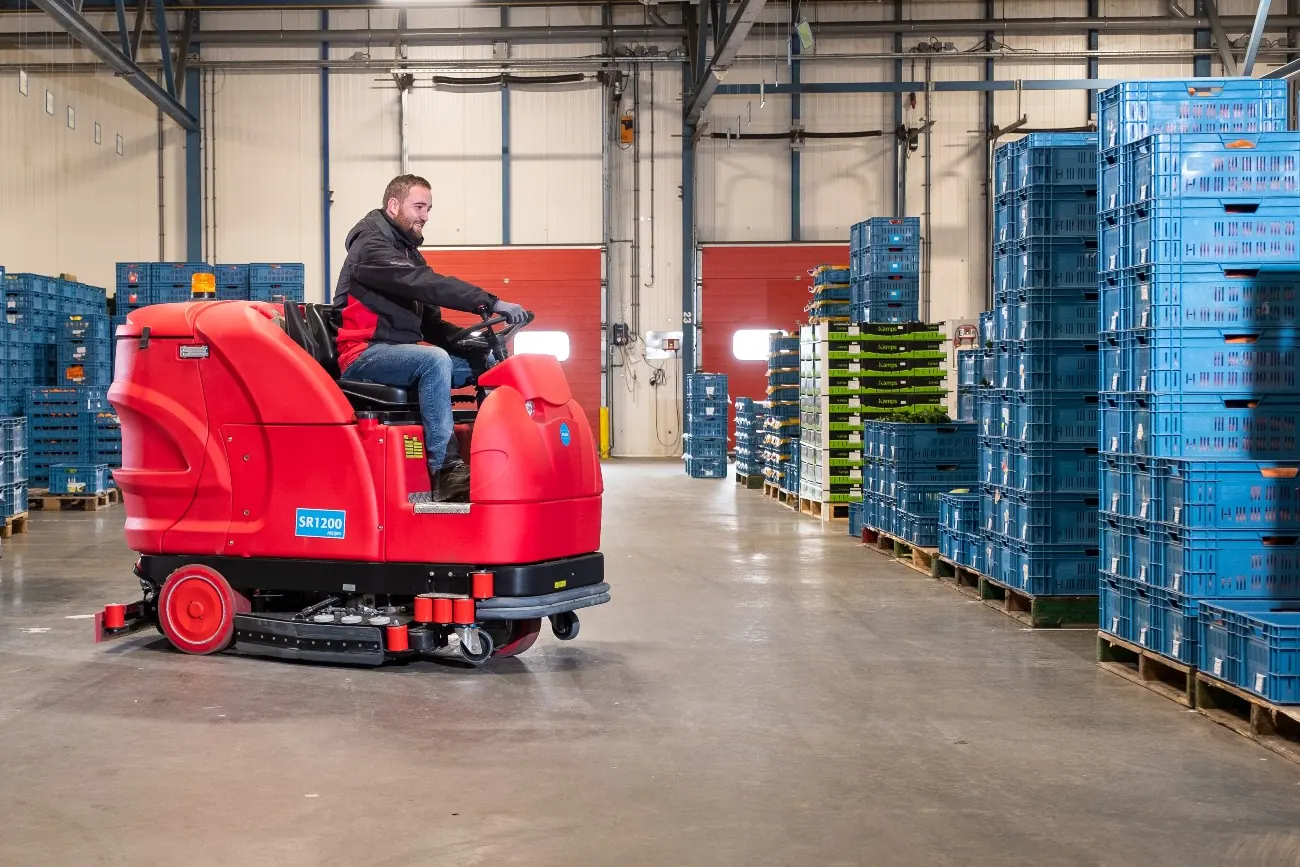 Medewerker rijdt met een Meijer SR1200 opzitschrobmachine door een distributiehal met blauwe kratten