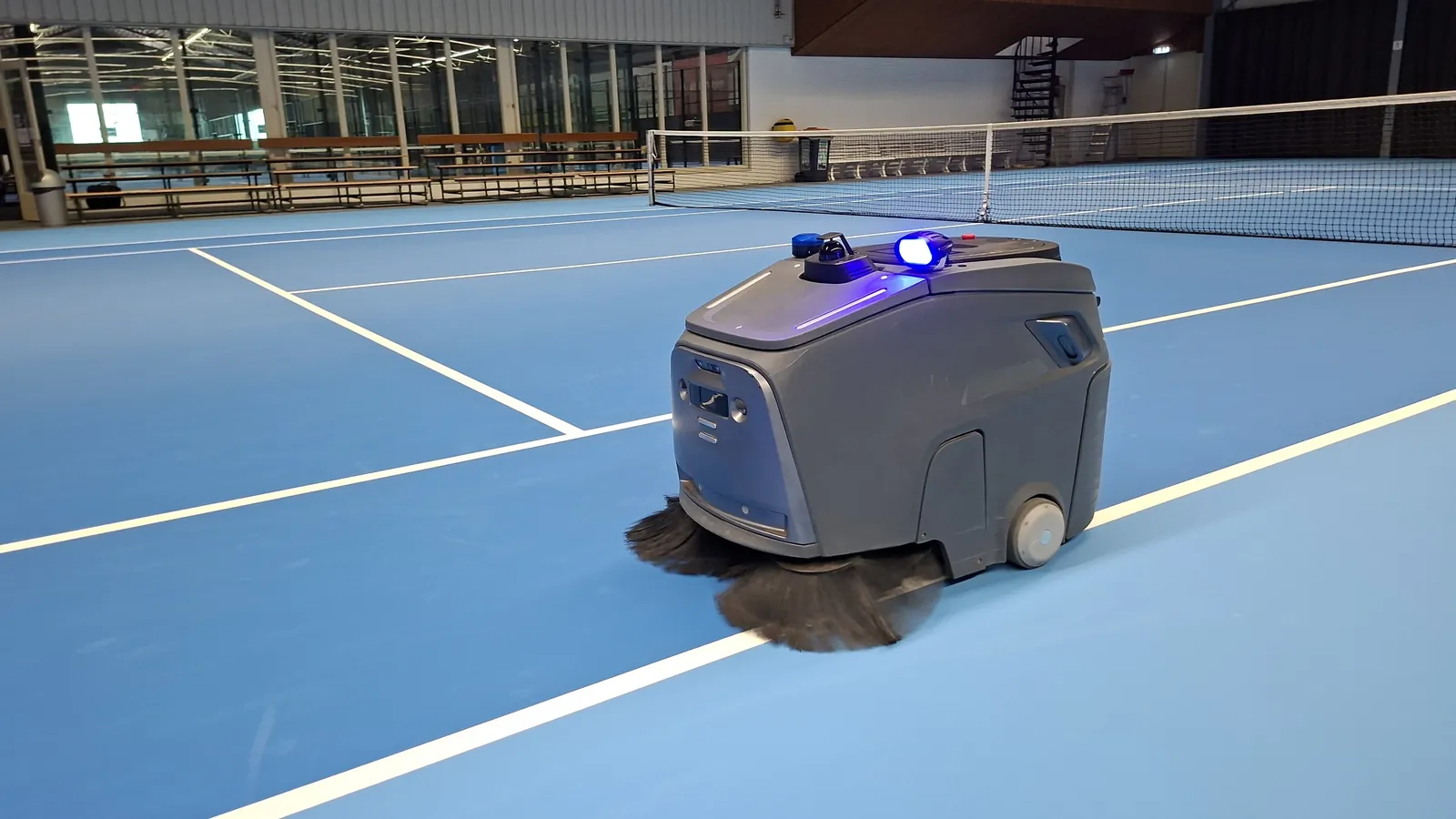 Gausium Beetle veegt een indoor tennisbaan, met draaiende zijborstels en de zwaailamp aan